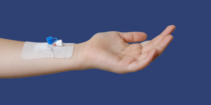 A close-up of a person's outstretched hand with a medical adhesive bandage on the wrist. The bandage has a small blue connector attached, indicating intravenous access. The background is a solid dark blue color.