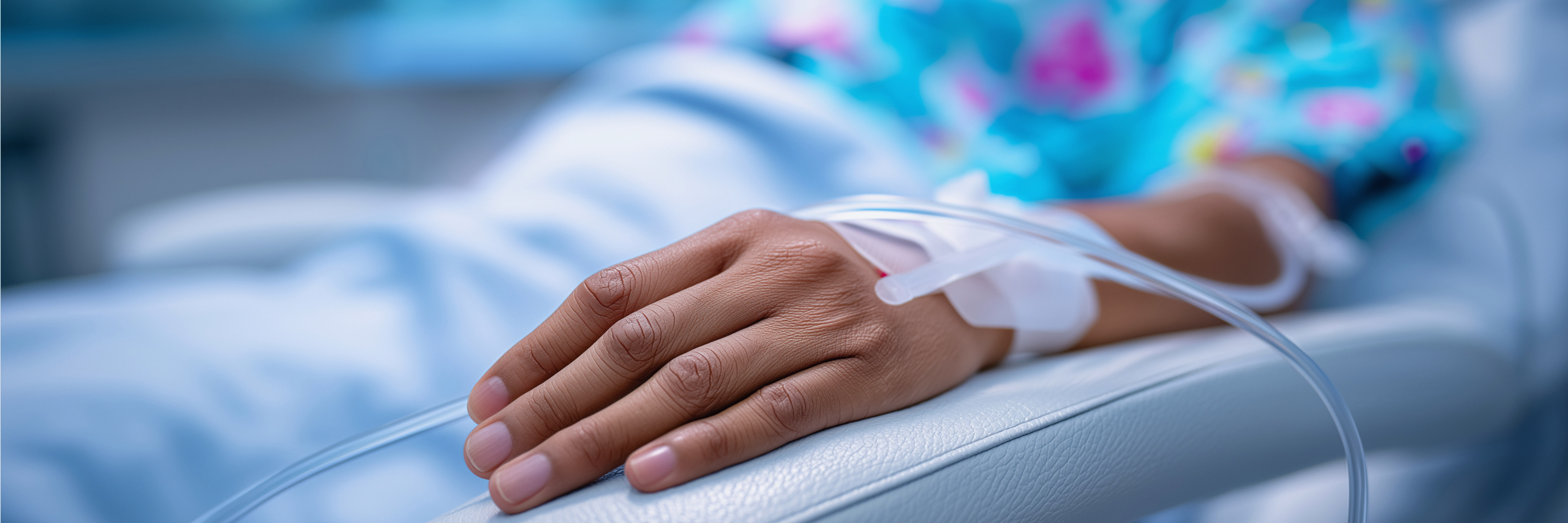 A close-up view of a patient's hand resting on a hospital bed, connected to an IV line. The hand is adorned in a colorful hospital gown, emphasizing a clinical setting. The background is softly blurred, suggesting a medical environment.