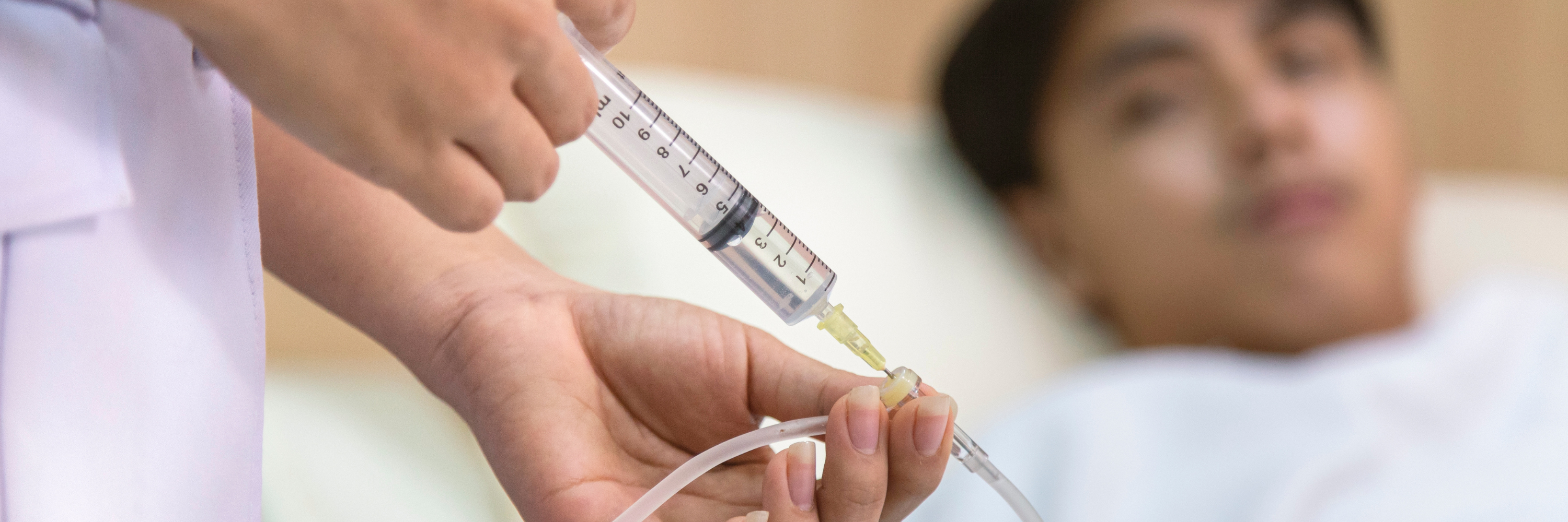 A healthcare professional is preparing to administer medicine using a syringe. In the background, a patient lies in a hospital bed, appearing calm as the procedure takes place. The focus is on the careful administration of treatment.